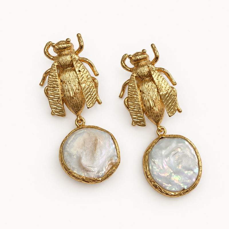 Coppia di orecchini con motivo insetto alato color oro e pendenti bianchi iridescenti su fondo chiaro.