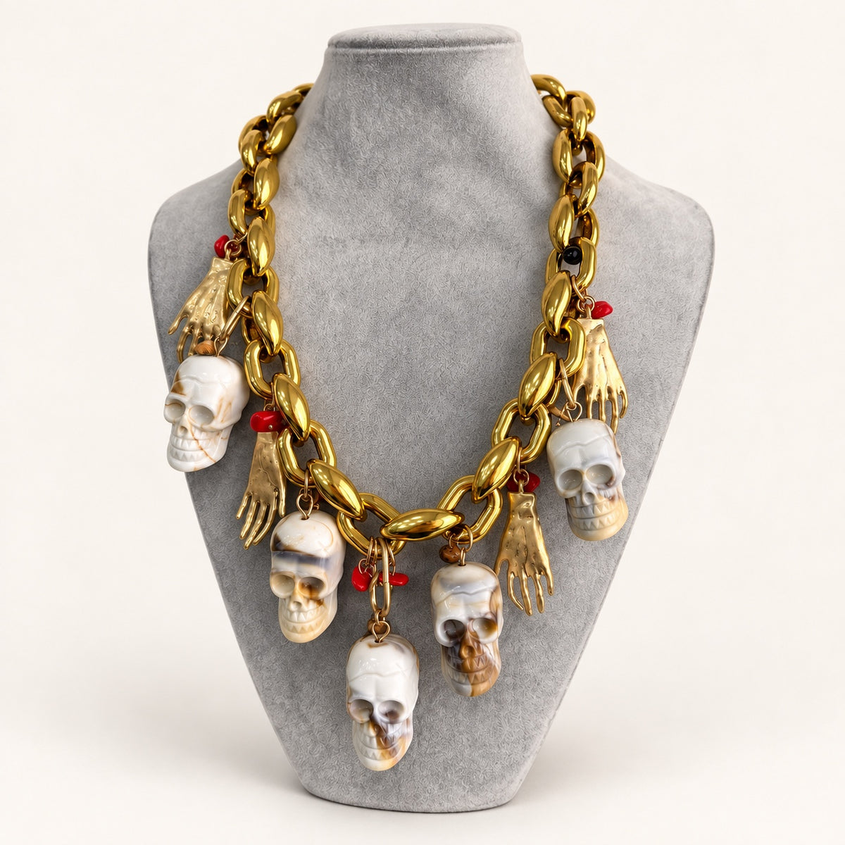 Collana a maglie grandi color oro vista di tre quarti, con pendenti a forma di teschio e mano su espositore grigio.